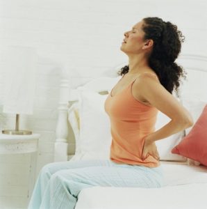 Woman sitting upright on bed holding back, eyes closed